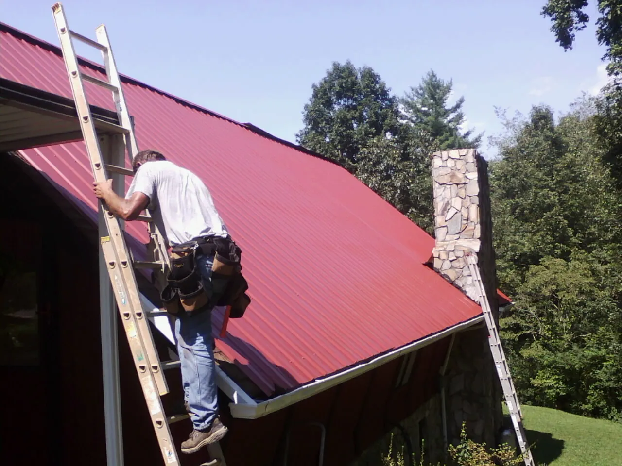 Roofer on a ladder beside a red metal roof and stone chimney — Soffit & Fascia Repair by Roofing Brothers in Long Island City